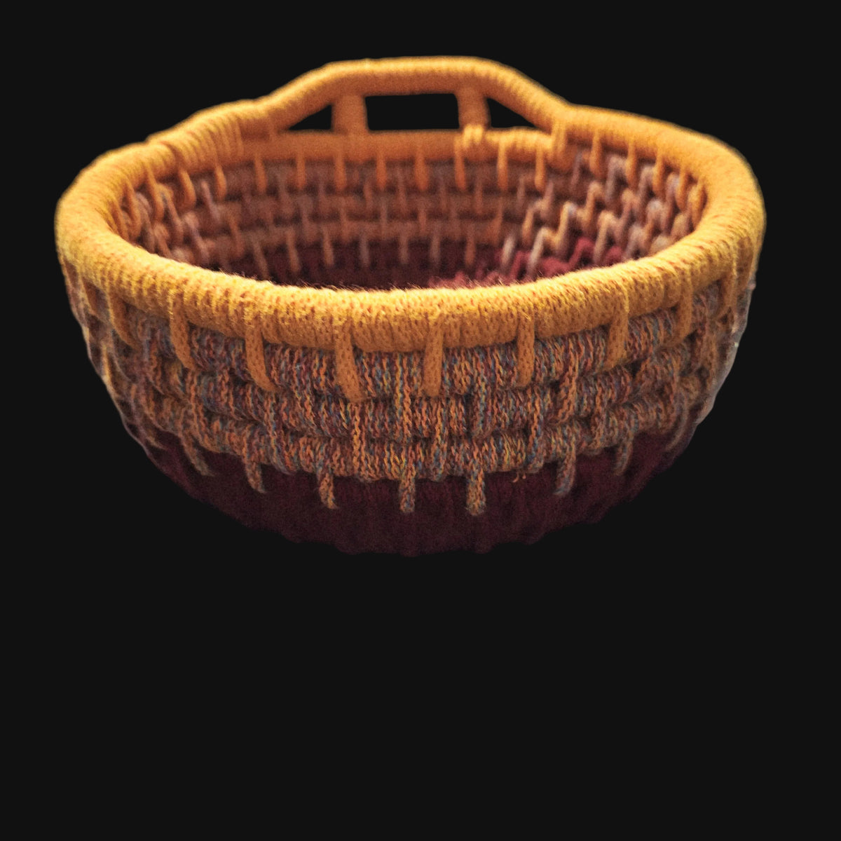 A coiled basket with a top handle, featuring a striped pattern in orange, terracotta, and burgundy. The center is  a yellow pumpkin design and the word 'Thankful' in burgundy script. 9x9x4 inches overall.