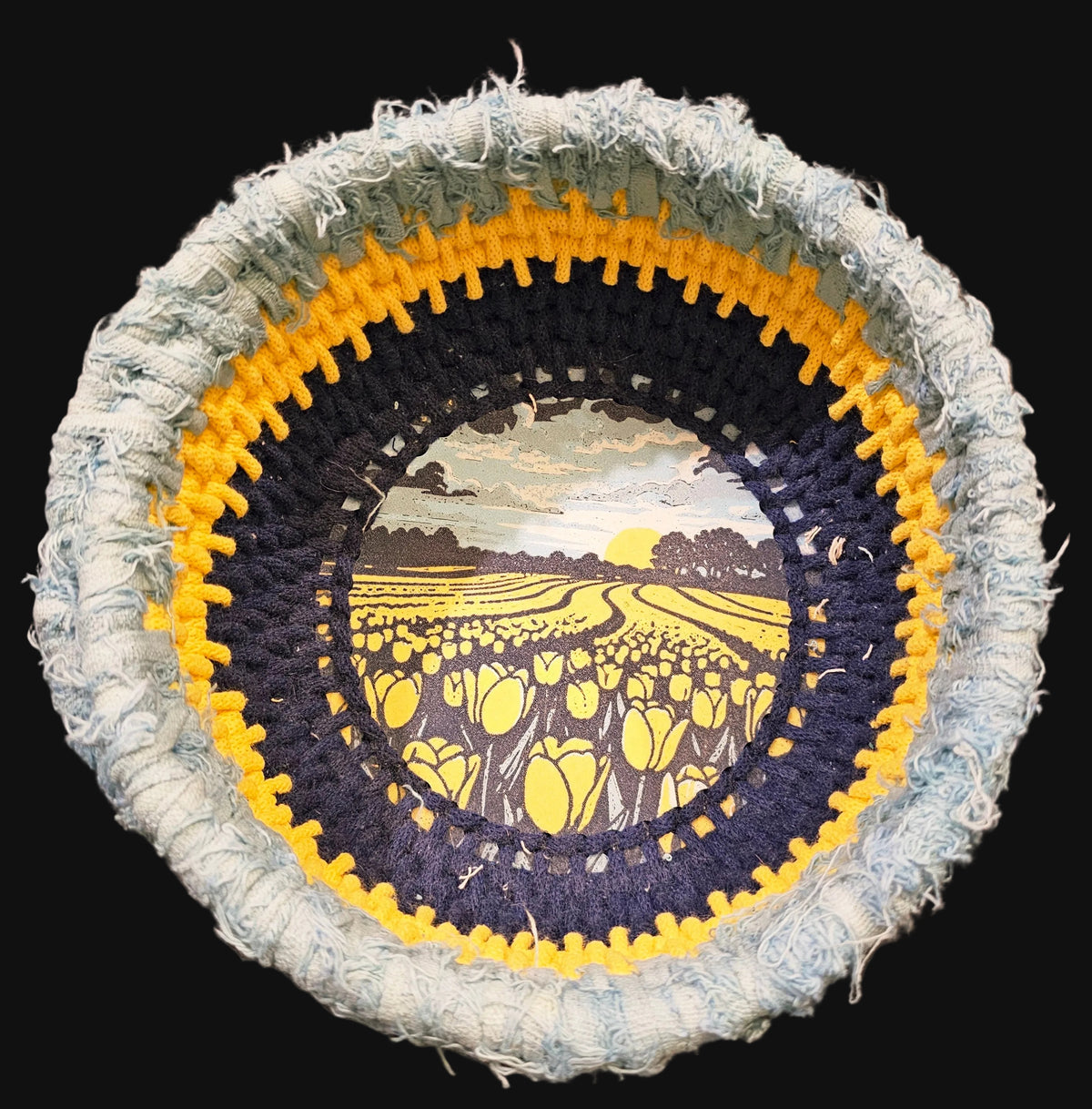A circular coiled basket with a rustic frayed outer edge, featuring concentric rings of light blue, yellow, and dark navy over a central printed illustration of a tulip field at sunset. 9x9x4 inches overall