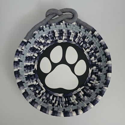 A coiled basket with a gray rope handle, featuring a black center with a white paw print design and a patterned border of navy, gray, and white woven strips.